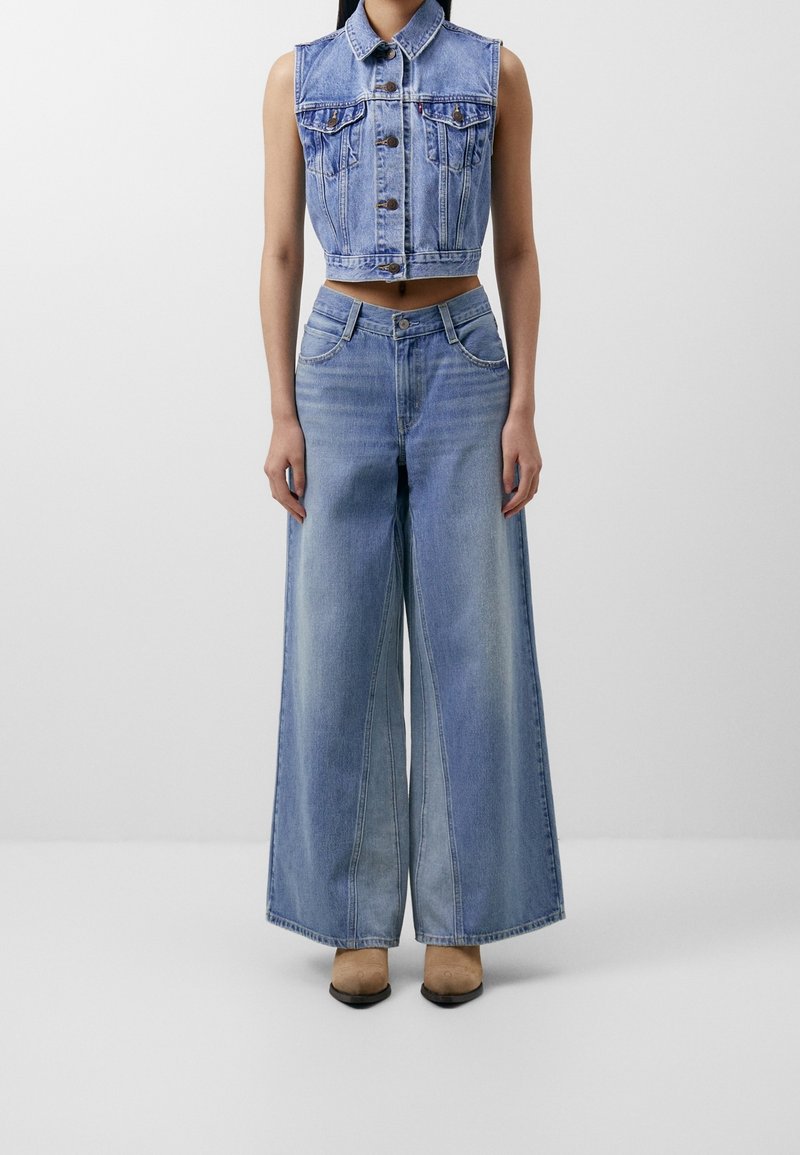 Femme portant un gilet en jean sans manches et court ainsi qu'un jean taille haute à jambes larges, avec des chaussures pointues beiges, sur fond blanc uni.