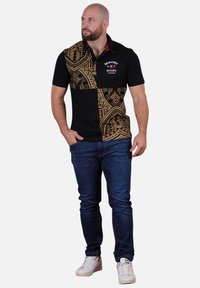 T-shirt polo noir avec un grand motif tribal brun sur un côté, manches courtes et un col avec des accents rouges. Le modèle est associé à un jean bleu.