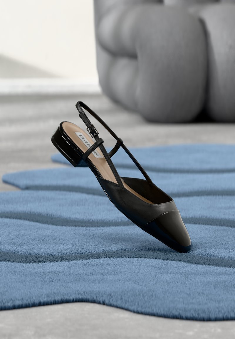 Fekete lakkbőr slingback cipő alacsony blokk sarokkal, texturált kék hullámos szőnyegen és szürke párnázott bútoron nyugszik.