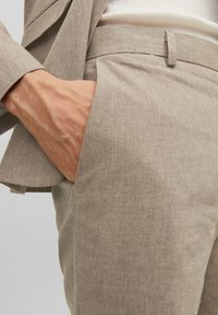 Pantaloni in lino beige con frontale piatto, tasche laterali e una texture liscia. Dettagli del tessuto sottili visibili, indossati con un blazer su misura.