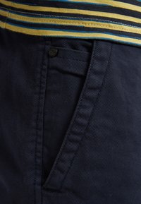 Námořnické modré bavlněné kalhoty s texturovanou tkaninou, které mají přední kapsu s černým knoflíkem, šité švy a viditelný pás se pruhy nahoře.