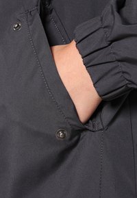 Czarna kurtka zbliżeniowym ujęciem otwartej kieszeni i zapięciem na zatrzask, z gromadzonymi mankietami i gładką fakturą.