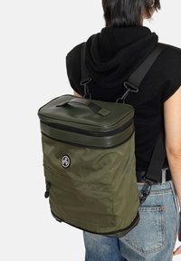 Sac à dos vert olive avec un design structuré, dotée d'un dessus zippé, de sangles tissées et d'une poche avant. Tissu texturé et détail du logo visibles.