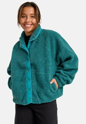 Donna sorridente che indossa una giacca in pile texturizzato color turchese con bottoni a pressione, mani nelle tasche e pantaloni neri su uno sfondo semplice.