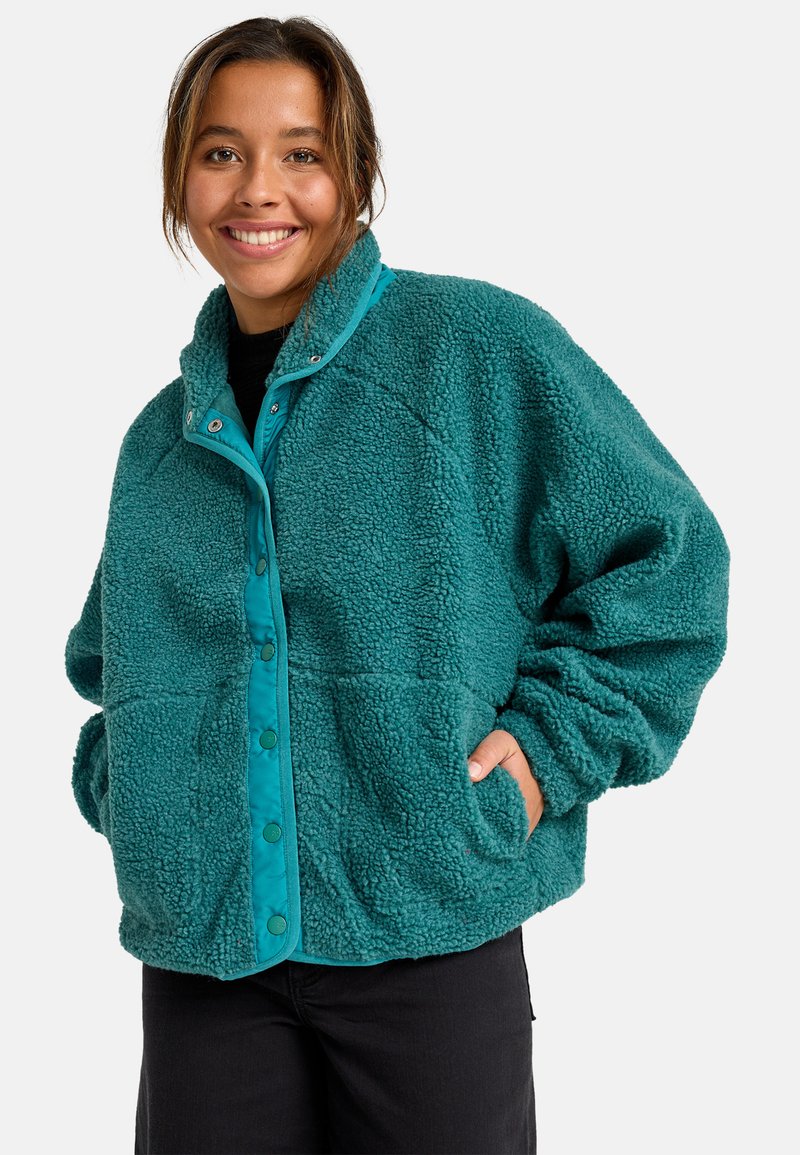 Lachende vrouw in een textuur fleece jack in teal met drukknoopsluitingen, handen in de zakken, en zwarte broek voor een effen achtergrond.