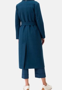 Long manteau ajusté en tissu bleu sarcelle foncé avec une taille ceinturée, présentant un dos droit et des détails à fentes latérales. Associé à un pantalon raccourci et des baskets.