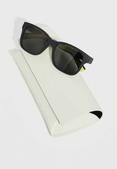 Μαύρα γυαλιά ηλίου με τετράγωνο σκελετό, ακουμπισμένα πάνω σε λευκή δερμάτινη θήκη με το ανάγλυφο όνομα της μάρκας, σε ανοιχτό φόντο.