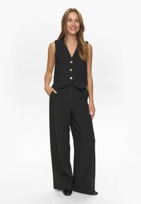 Chaleco negro sin mangas con cuello de muesca, que presenta un cierre de botón frontal y dos bolsillos laterales; combinado con pantalones holgados negros.