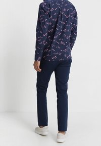 Dunkelblaues Hemd mit rosa Blumenmuster, zugeknöpft, kombiniert mit dunkelblauen Hosen. Die Person trägt weiße Turnschuhe. Ansicht von hinten.