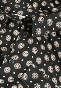 Schwarzes Hemd aus Stoff mit einem wiederholten kreisförmigen Muster in Weiß und Orange, versehen mit kleinen weißen Punkten. Sechs schwarze Knöpfe sichtbar.