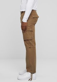 Pantalons cargo marron en coton avec poches latérales, dotés d'une jambe effilée et d'ourlets ajustables. Portés avec des baskets blanches.