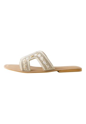 Füße in beigen Sandalen mit quadratischer Spitze und weißen Perlen- und Perlenverzierungen unter einem schwarzen transparenten Rock.