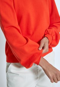 Orange sweatshirt with wide, gathered sleeves. Made from soft cotton blend fabric. Worn over light-colored pants, showcasing a relaxed fit.