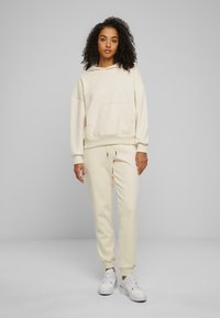 Sweat-shirt beige à capuche avec une poche frontale, associé à un pantalon de jogger assorti. Les deux sont fabriqués en tissu doux, avec des poignets élastiques et une taille à cordon de serrage.