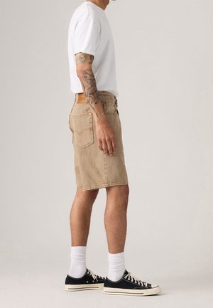 Mand stående i beige denimshorts, hvid t-shirt, hvide sokker og sorte lave sneakers, med tatoveret arm synlig fra siden.
