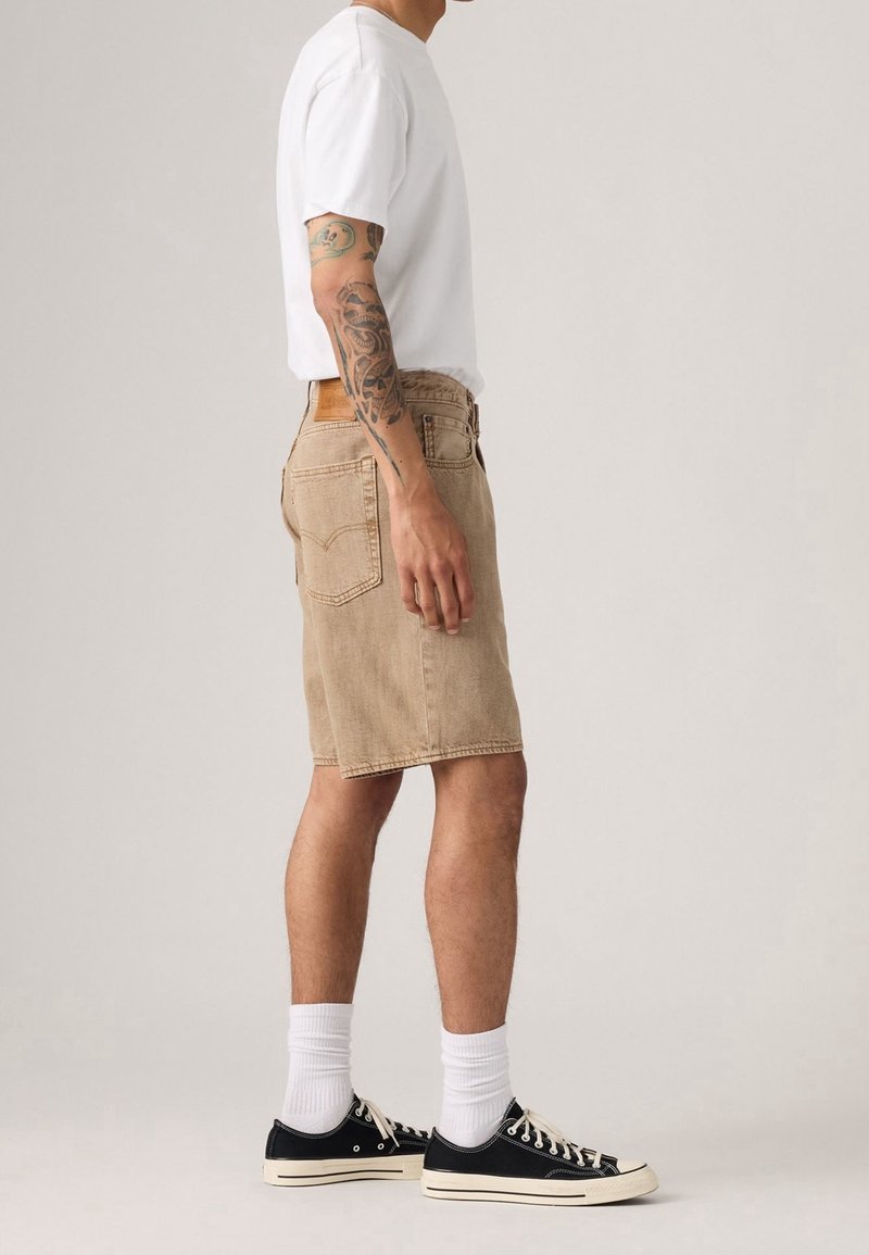 Homme debout en short en jean beige, t-shirt blanc, chaussettes blanches et baskets basses noires, avec un bras tatoué visible de profil.