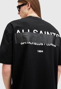 Svart bomull T-skjorte med et stort hvitt trykt logo på baksiden, med 'ALLSAINTS SPITALFIELDS / LONDON' og '1994' i hvit tekst.