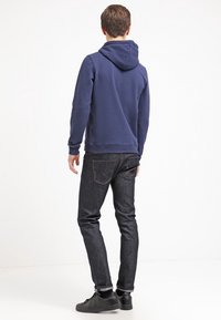 Felpa blu navy con tasca a canguro frontale, tessuto testurizzato e cappuccio regolabile, abbinata a jeans scuri e sneaker nere.
