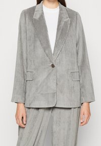 Blazer di velluto a coste grigio con rever a punta, chiusura a un bottone e due tasche frontali. Presenta una texture a coste verticali.