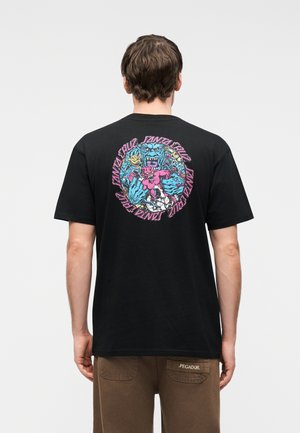 Άνδρας που φορά μαύρο μπλουζάκι με πολύχρωμο γραφικό από μπλε και ροζ τέρατα και επαναλαμβανόμενο κείμενο «Santa Cruz» στην πλάτη, σε συνδυασμό με καφέ παντελόνι.