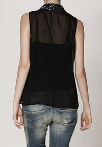 Top senza maniche nero con pannello posteriore trasparente, colletto dettagliato con accenti metallici, abbinato a jeans blu sbiaditi con tasche posteriori.