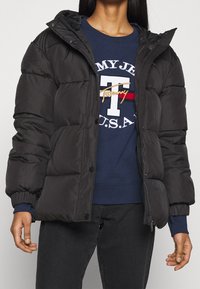 Czarna kurtka puchowa z pikowanym wzorem, ściągaczami przy rękawach i zamkiem na przodzie. Noszona na granatowym swetrze z nadrukiem logo.