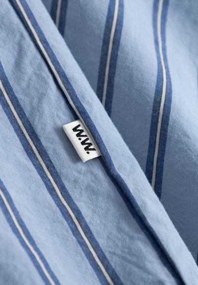 Tela azul claro con rayas verticales oscuras y blancas y una pequeña etiqueta de ropa blanca con un logo negro de estilo manuscrito.