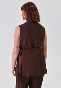 Persona con capelli castano ondulati di lunghezza media che indossa un top senza maniche marrone scuro con una cintura annodata, vista da dietro su uno sfondo neutro.