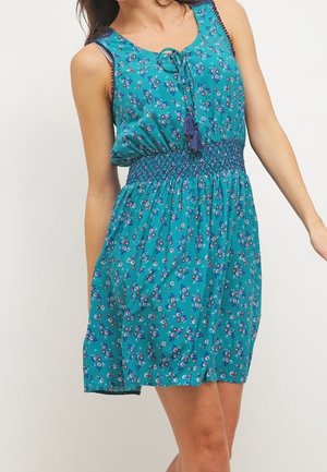 Robe sans manches turquoise à fleurs avec taille smockée et lien à pampilles au niveau du col portée par une personne debout.