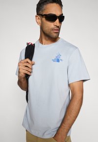 Ανοιχτό μπλε βαμβακερό μπλουζάκι με γραφικό λογότυπο σε μπλε χρώμα. Το μοντέλο κρατάει ένα μαύρο σακίδιο πλάτης με κόκκινες λεπτομέρειες σε έναν ώμο.