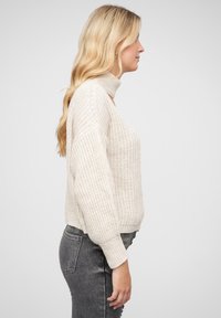 Cream-colored knit sweater with a high collar, loose fit, and textured pattern. Ribbed cuffs and hem, worn over gray jeans.