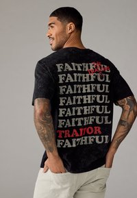 Zwarte katoenen t-shirt met een gestructureerde afwerking en een gedurfde tekstontwerp op de achterkant, met herhaalde woorden "FAITHFUL" en "TRAITOR" in grijs en rood.