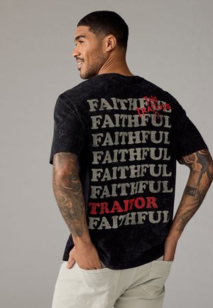 Svart bomull t-shirt med en strukturerad finish och fet textdesign på baksidan, med upprepade ord "TROGEN" och "FÖRRAD" i grått och rött.