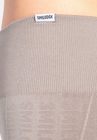 Beige ribbet talje på leggings med "SMILODOX" etiket. Blød tekstur med subtilt mønster, der smalner ud til et glat ben.