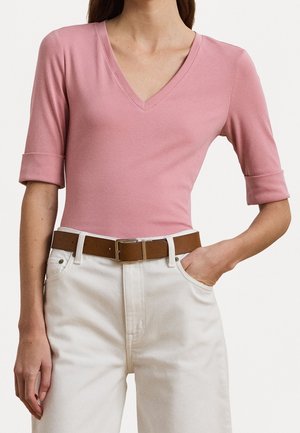 Femme portant un haut rose à col en V avec manches retroussées aux coudes, pantalon blanc taille haute et ceinture en cuir marron, la main dans la poche.