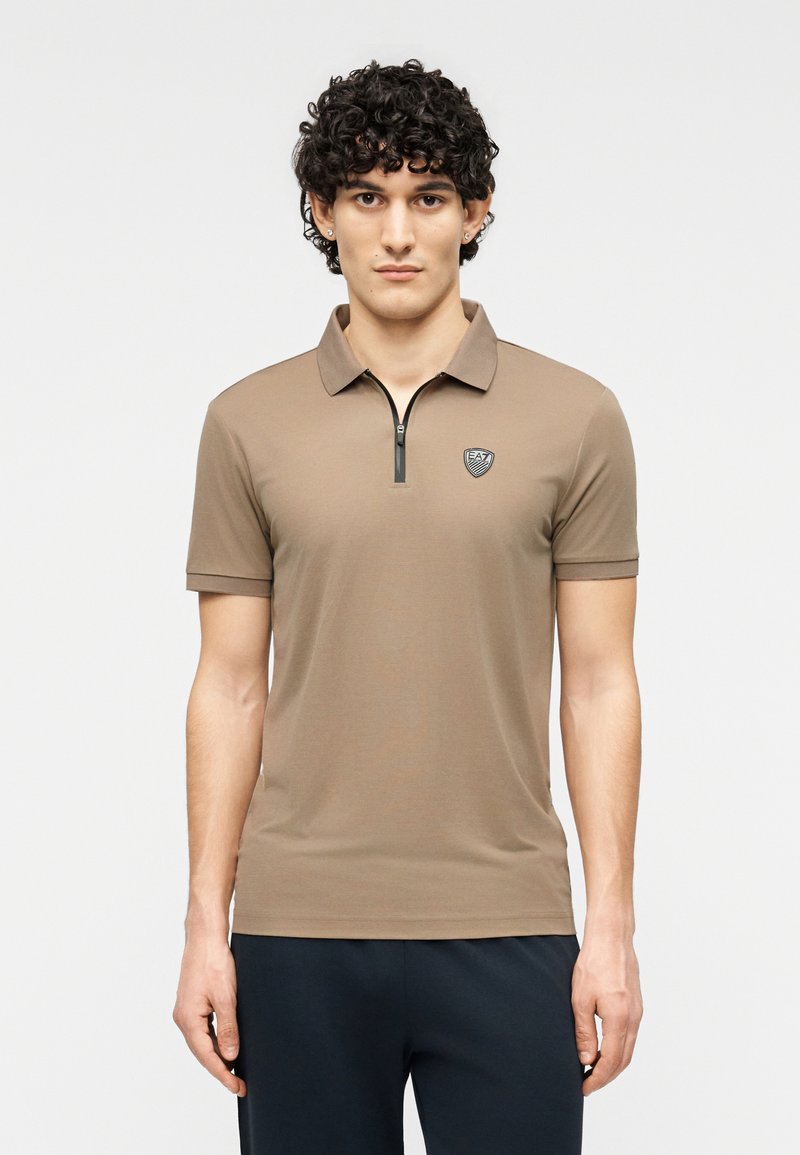 Καφέ πουκάμισο πόλο με κοντά μανίκια, σχεδιασμένο με φερμουάρ και μικρό λογότυπο στο αριστερό στήθος. Λεία υφή και εφαρμοστή σιλουέτα.