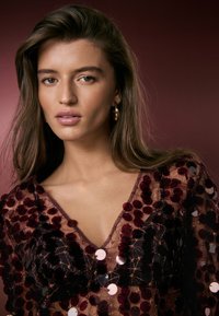 Sheer dress with deep V-neck, adorned with dark sequins and a subtle pattern, featuring a lightweight texture against a burgundy background.
