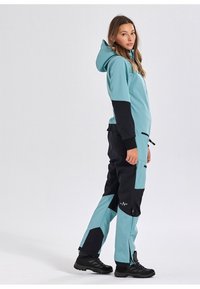 Een twee- kleurige softshell overall in lichtblauw en zwart, met een capuchon, ritssluitingzakken, en zwarte accenten op de mouwen en benen. Heeft een aangesloten pasvorm.