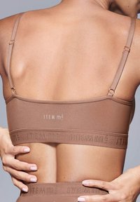 Reggiseno wireless marrone con spalline regolabili. Presenta il logo e una fascia testurizzata sul fondo. Tessuto liscio con un design minimalista.