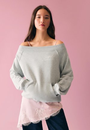 Junge Frau mit langen braunen Haaren trägt ein schulterfreies graues Sweatshirt mit dem Text "Cupid Loves Me", ein rosa, mit Spitze besetztes Oberteil und dunkle Hosen vor einer pinken Wand.