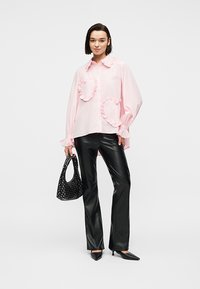 Γυναίκα που φοράει ροζ μπλούζα με βολάν σε σχήμα καρδιάς, μαύρο δερμάτινο παντελόνι, μαύρα τακούνια kitten, κρατώντας μία μαύρη τσάντα με τρουκς.