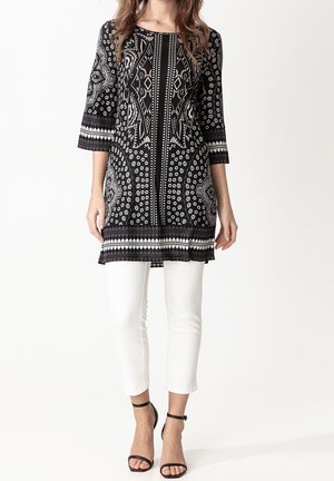 Vrouw draagt een zwart-wit patroon tuniek met driekwart mouwen, witte cropped broek en zwarte enkellaarzen met riempje.