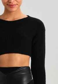 Schwarzer gerippter Crop-Pullover mit langen Ärmeln, der einen weiten Ausschnitt und eine weiche Textur aufweist. Kombiniert mit hoch taillierten schwarzen Lederhosen.