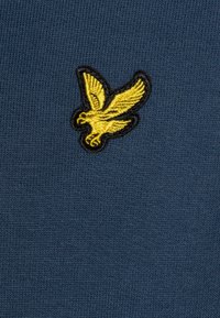 Tessuto a maglia navy con logo di un uccello ricamato giallo bordato di nero. La texture è liscia con dettagli di cucitura ben definiti.