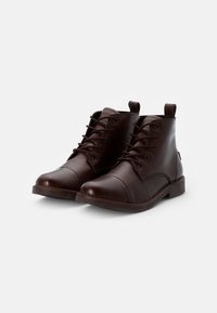 Par de botas de cuero marrón oscuro con cordones hasta el tobillo, con tiradores, punteras redondeadas y tacones bajos apilados sobre un fondo blanco.