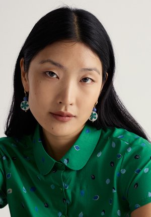 Groen overhemd met een bladpatroon. Het model draagt ronde oorbellen met een blauw kraaltje en een bloemmotief in groen en wit.