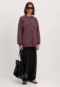 Übergröße, weinroter Pullover mit gerippten Bündchen, kombiniert mit schwarzen weiten Hosen und einer glänzenden schwarzen Tragetasche. Trägt außerdem dunkle Sonnenbrille.