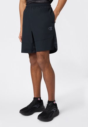 Uomo con pantaloncini sportivi neri e scarpe da ginnastica nere abbinate, in piedi contro uno sfondo chiaro uniforme.