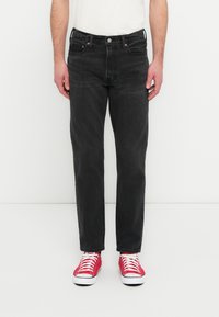 Μαύρο τζιν από denim με ίσια γραμμή στα πόδια, εμφανείς ραφές, πέντε τσέπες και κούμπωμα με κουμπιά. Φοριέται με κόκκινα πάνινα παπούτσια.