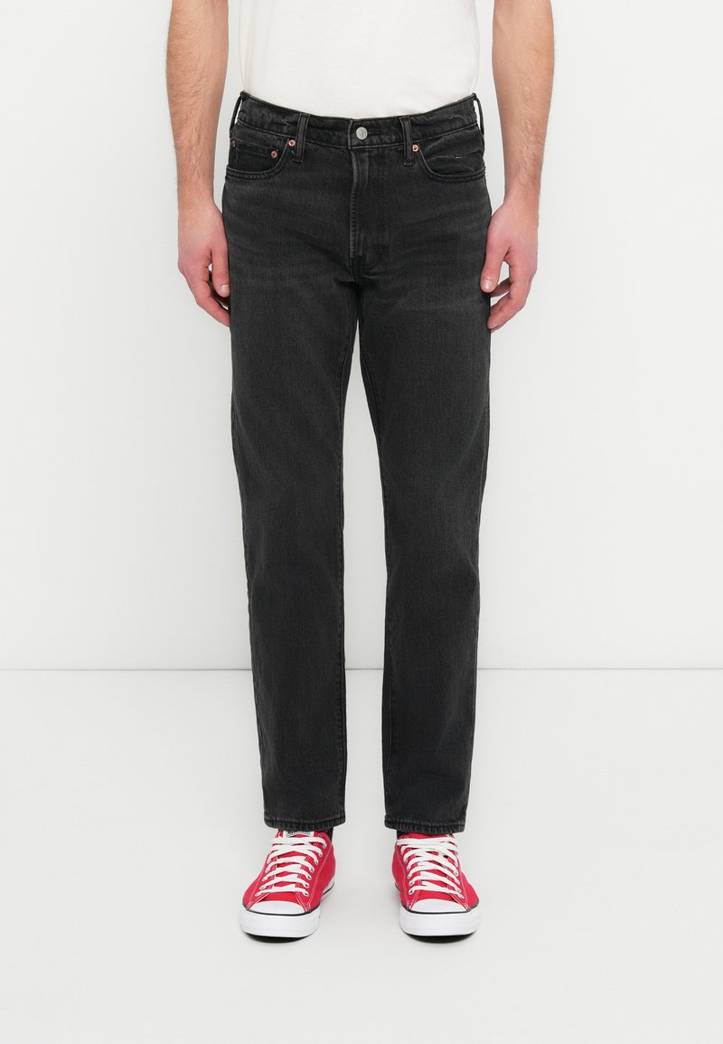 Μαύρο τζιν από denim με ίσια γραμμή στα πόδια, εμφανείς ραφές, πέντε τσέπες και κούμπωμα με κουμπιά. Φοριέται με κόκκινα πάνινα παπούτσια.